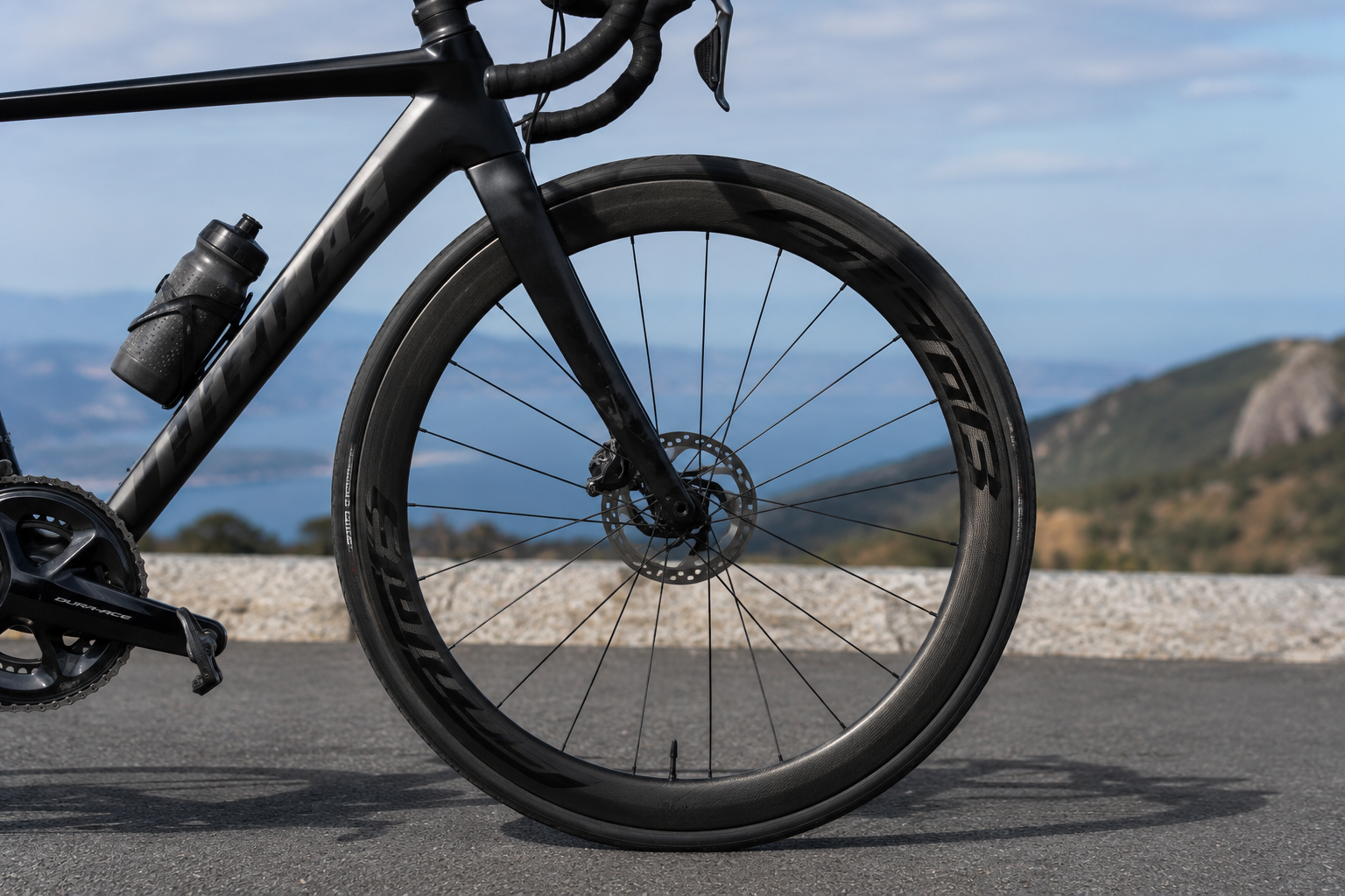 Close-up of a high-performance road bicycle featuring deep-section carbon wheels on a paved road, highlighting aerodynamic design and premium wheelset construction against a scenic outdoor backdrop