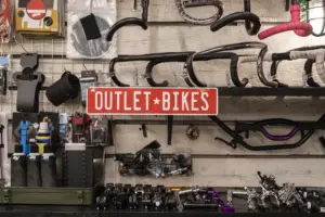 Interior view of a bike shop workshop wall featuring handlebars, bike parts, tools, and a red "OUTLET BIKES" sign, illustrating custom bicycle fitting and repair services in West LA.