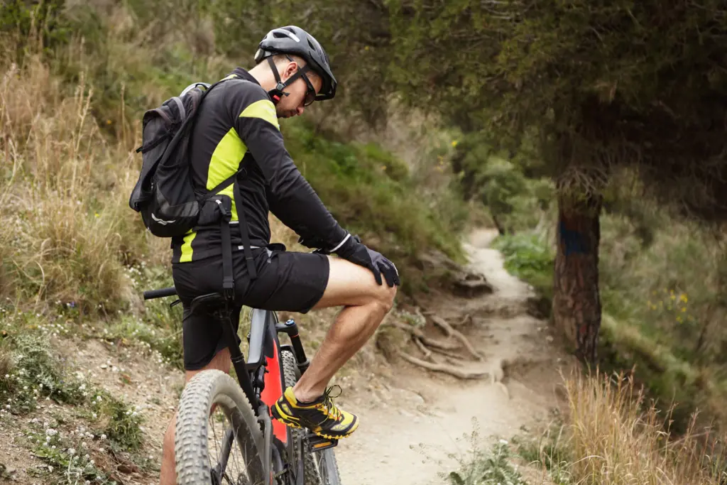 A mountain biker in a helmet, backpack, and black-and-neon-yellow gear sits on his bike on a dirt trail, wearing yellow and black shoes that illustrate mountain bike shoe fit and cleat setup for clipless vs flat pedals.