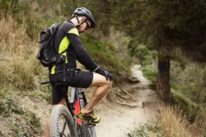 A mountain biker in a helmet, backpack, and black-and-neon-yellow gear sits on his bike on a dirt trail, wearing yellow and black shoes that illustrate mountain bike shoe fit and cleat setup for clipless vs flat pedals.